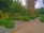 A well-maintained, lush green park with various trees and shrubs lining a paved walkway. The vibrant green foliage is abundant, and the tall trees provide a canopy overhead. There are stone planters and a pathway leading through the park, and the sky is partially visible.