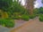 A well-maintained, lush green park with various trees and shrubs lining a paved walkway. The vibrant green foliage is abundant, and the tall trees provide a canopy overhead. There are stone planters and a pathway leading through the park, and the sky is partially visible.