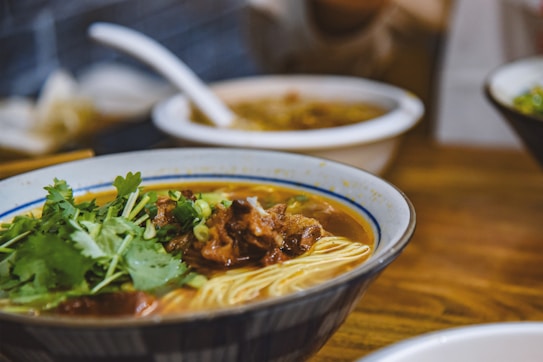 A bowl of noodle soup garnished with fresh cilantro and green onions, topped with pieces of braised meat. The soup appears rich and savory, with another bowl of food blurred in the background on a wooden table.