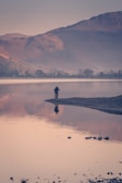 A soft-focus photograph of a solitary figure standing by a foggy lake at dawn, the light diffused and gentle.