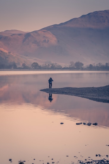 A lone figure stands on a small land strip by the edge of a serene lake, tripod set up, capturing the expansive landscape. Mist envelops the distant mountains, bathed in soft, warm light of either dawn or dusk, reflecting in the calm water.