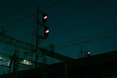 A nighttime scene featuring a complex network of overhead power lines and a signal light displaying red. The scene is dark, illuminated by faint artificial lights in the background.