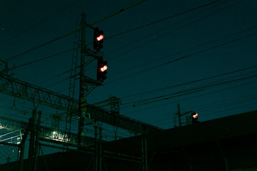 A nighttime scene featuring a complex network of overhead power lines and a signal light displaying red. The scene is dark, illuminated by faint artificial lights in the background.