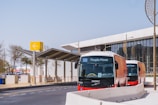 Two modern People Mover buses are parked near a station with a contemporary design. A large yellow sign with an arrow is visible, along with a building featuring glass windows and decorative round structures.