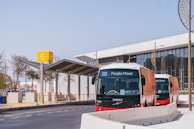 Two modern People Mover buses are parked near a station with a contemporary design. A large yellow sign with an arrow is visible, along with a building featuring glass windows and decorative round structures.
