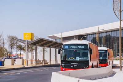 Two modern People Mover buses are parked near a station with a contemporary design. A large yellow sign with an arrow is visible, along with a building featuring glass windows and decorative round structures.
