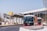 Two modern People Mover buses are parked near a station with a contemporary design. A large yellow sign with an arrow is visible, along with a building featuring glass windows and decorative round structures.