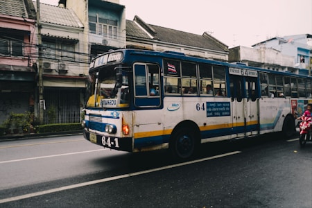 A vintage public bus driving down a city street, with old buildings in the background. The bus is painted in white and blue with yellow accents, featuring route number 64. Passengers are visible through the windows. A motorbike passes by on the right side of the image.