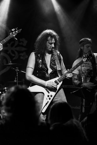 A rock guitarist is performing on stage, wearing a leather vest and holding a Flying V electric guitar. Spotlights illuminate the scene, casting beams of light. Another musician is partially visible, playing a guitar with a focused expression.