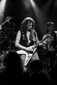 A rock guitarist is performing on stage, wearing a leather vest and holding a Flying V electric guitar. Spotlights illuminate the scene, casting beams of light. Another musician is partially visible, playing a guitar with a focused expression.