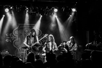 A black and white photograph of a live music performance featuring a rock band, with three musicians playing guitars on stage. Smoke and dramatic lighting create a dynamic and intense atmosphere, while an audience is visible in the foreground, some holding phones capturing the event.