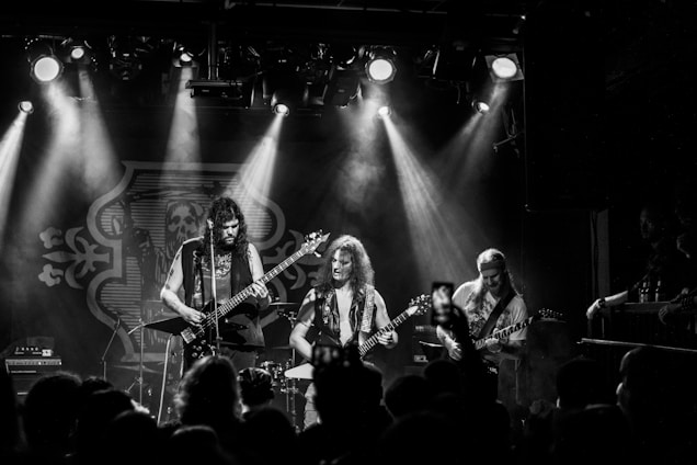 A black and white photograph of a live music performance featuring a rock band, with three musicians playing guitars on stage. Smoke and dramatic lighting create a dynamic and intense atmosphere, while an audience is visible in the foreground, some holding phones capturing the event.