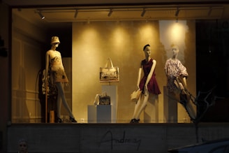 A fashion store window display showcases three mannequins dressed in stylish outfits. The left mannequin wears a printed dress and bucket hat, while the central one dons a sleeveless burgundy dress, clutching a handbag. The right mannequin is dressed in a patterned, voluminous-sleeved top and leans against a stand. A handbag is prominently displayed on a pedestal in front.