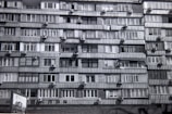 A multi-story residential building features numerous apartments with balconies. Each unit is fitted with air conditioning units attached below the windows. The facade exhibits a uniform pattern of windows and concrete, creating a repetitive architectural style.