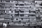A multi-story residential building features numerous apartments with balconies. Each unit is fitted with air conditioning units attached below the windows. The facade exhibits a uniform pattern of windows and concrete, creating a repetitive architectural style.