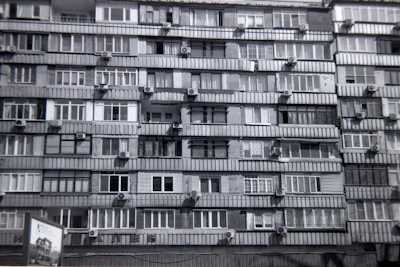 A multi-story residential building features numerous apartments with balconies. Each unit is fitted with air conditioning units attached below the windows. The facade exhibits a uniform pattern of windows and concrete, creating a repetitive architectural style.