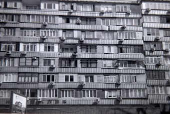 A multi-story residential building features numerous apartments with balconies. Each unit is fitted with air conditioning units attached below the windows. The facade exhibits a uniform pattern of windows and concrete, creating a repetitive architectural style.
