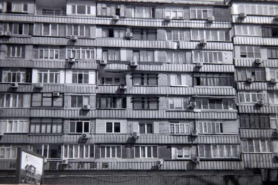 A multi-story residential building features numerous apartments with balconies. Each unit is fitted with air conditioning units attached below the windows. The facade exhibits a uniform pattern of windows and concrete, creating a repetitive architectural style.