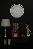 A wall-mounted clock with a silver frame and black numbers is displayed against a dark background. Below the clock, there is a tabletop arrangement featuring a lamp with a beige shade, a vintage red gumball machine partially filled with colorful candies, a ceramic mug, a wooden artist's mannequin in a dynamic pose, and a small white cubic device. Glass jars filled with shells and small items are placed on the lower shelf.