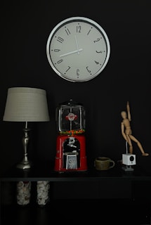 A wall-mounted clock with a silver frame and black numbers is displayed against a dark background. Below the clock, there is a tabletop arrangement featuring a lamp with a beige shade, a vintage red gumball machine partially filled with colorful candies, a ceramic mug, a wooden artist's mannequin in a dynamic pose, and a small white cubic device. Glass jars filled with shells and small items are placed on the lower shelf.