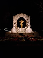 A brightly illuminated golden statue of a violinist stands prominently within a decorative stone arch. Surrounding the figure are intricate carvings depicting figures in dynamic poses. In the foreground, a variety of flowers, including many red tulips, create a vibrant border. The scene is set against a dark, nighttime backdrop, with the statue and arch lit dramatically.