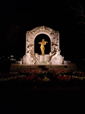A brightly illuminated golden statue of a violinist stands prominently within a decorative stone arch. Surrounding the figure are intricate carvings depicting figures in dynamic poses. In the foreground, a variety of flowers, including many red tulips, create a vibrant border. The scene is set against a dark, nighttime backdrop, with the statue and arch lit dramatically.