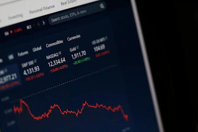 A computer screen displays a financial dashboard with stock market data, including indices like S&P 500 and NASDAQ, along with commodities like gold prices. A red line graph shows a declining trend, indicating negative market performance.
