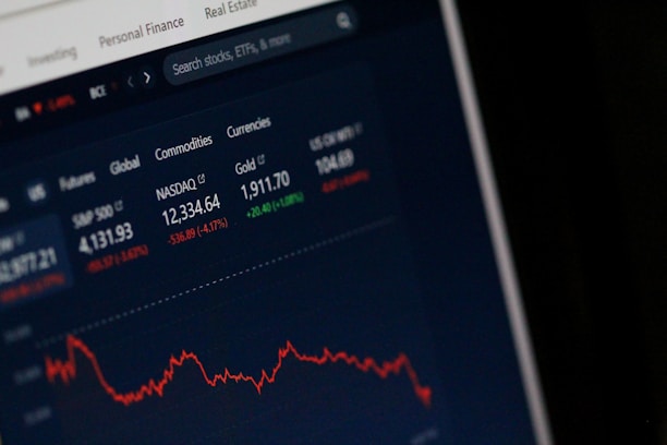 A computer screen displays a financial dashboard with stock market data, including indices like S&P 500 and NASDAQ, along with commodities like gold prices. A red line graph shows a declining trend, indicating negative market performance.