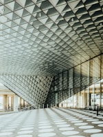 A modern architectural space features a geometric ceiling made of triangular patterns, creating an intriguing visual effect. The floor is covered in large white circular patterns contrasting with the dark background. Sunlight streams through the large glass windows on the side, illuminating the area.