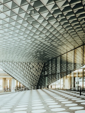 A modern architectural space features a geometric ceiling made of triangular patterns, creating an intriguing visual effect. The floor is covered in large white circular patterns contrasting with the dark background. Sunlight streams through the large glass windows on the side, illuminating the area.