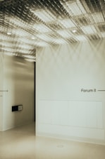 A minimalist interior design featuring a textured ceiling with a grid pattern casting shadows on the walls. The walls are smooth and painted in a light, neutral color. On the right side, there is text reading 'Forum II' indicating a direction. The lighting is soft, adding to the calm ambiance.