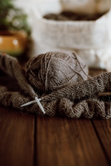 A ball of brown yarn is placed on a wooden surface next to knitting needles. The yarn is partially unwound and appears to be in the process of knitting. In the blurred background, there is a plant in a pot and a white bag partially in view.