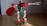 A toy constructed from interlocking plastic bricks resembling a robot with red, green, and white parts. It features a gear-shaped shield in one hand and a mechanical arm extended outward. The floor is wooden, and the background is a plain, grayish wall.