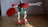 A toy constructed from interlocking plastic bricks resembling a robot with red, green, and white parts. It features a gear-shaped shield in one hand and a mechanical arm extended outward. The floor is wooden, and the background is a plain, grayish wall.