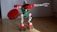 A toy constructed from interlocking plastic bricks resembling a robot with red, green, and white parts. It features a gear-shaped shield in one hand and a mechanical arm extended outward. The floor is wooden, and the background is a plain, grayish wall.
