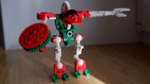 A toy constructed from interlocking plastic bricks resembling a robot with red, green, and white parts. It features a gear-shaped shield in one hand and a mechanical arm extended outward. The floor is wooden, and the background is a plain, grayish wall.