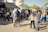 A bustling outdoor market scene with multiple stalls covered by colorful tents. People walk along a street lined with vendors, some browsing and others engaging in conversation. A man stands with a bicycle, raising his hand, and various goods are displayed on tables. The atmosphere is lively and communal, with a sunny day casting light and shadows.