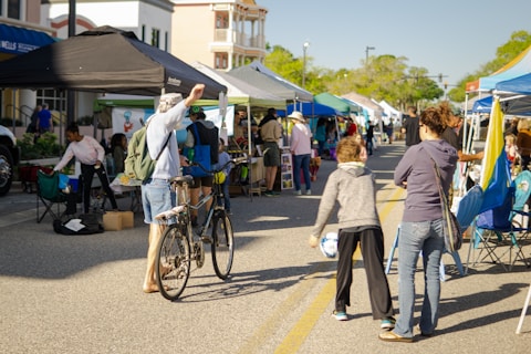 A bustling outdoor market scene with multiple stalls covered by colorful tents. People walk along a street lined with vendors, some browsing and others engaging in conversation. A man stands with a bicycle, raising his hand, and various goods are displayed on tables. The atmosphere is lively and communal, with a sunny day casting light and shadows.