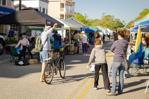 A bustling outdoor market scene with multiple stalls covered by colorful tents. People walk along a street lined with vendors, some browsing and others engaging in conversation. A man stands with a bicycle, raising his hand, and various goods are displayed on tables. The atmosphere is lively and communal, with a sunny day casting light and shadows.
