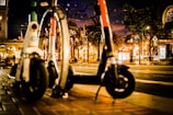 Two electric scooters are parked on a city sidewalk at night. There are palm trees and a clock tower in the background, and the street is illuminated by various lights and signs.