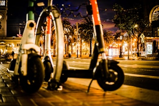 Two electric scooters are parked on a city sidewalk at night. There are palm trees and a clock tower in the background, and the street is illuminated by various lights and signs.