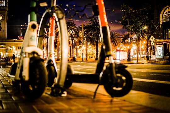 Two electric scooters are parked on a city sidewalk at night. There are palm trees and a clock tower in the background, and the street is illuminated by various lights and signs.