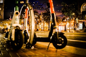 Two electric scooters are parked on a city sidewalk at night. There are palm trees and a clock tower in the background, and the street is illuminated by various lights and signs.