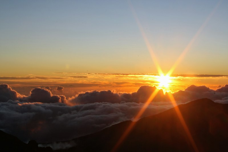 Sun breaking above a thick layer of cloud with a dark volcanic ridge silhouetted in the foreground