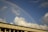 A vibrant rainbow arcs across a clear blue sky above a large building with a row of windows and a dark green roof. Fluffy white clouds are scattered around, and a faint view of the moon is also visible in the sky.