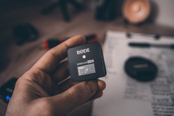 A hand is holding a RØDE Wireless GO II audio transmitter. The device is small, black, and square-shaped, with a digital display showing status indicators. In the background, there is a blurred clipboard with handwritten notes and a camera lens cap.