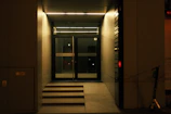 An entrance to a building featuring glass doors with a stepped access way and illuminated by overhead lighting. The area is bordered by smooth panels, and a scooter is parked to the side.