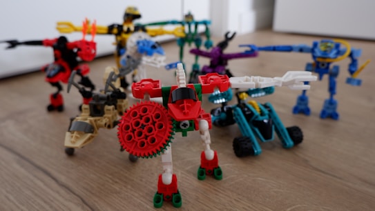 A group of colorful, articulated robot figures made from interlocking plastic pieces is arranged on a wooden floor. Each figure features distinct colors and shapes, with some resembling vehicles or animals equipped with mechanical limbs. The scene is playful and dynamic, showcasing a variety of textures and designs.
