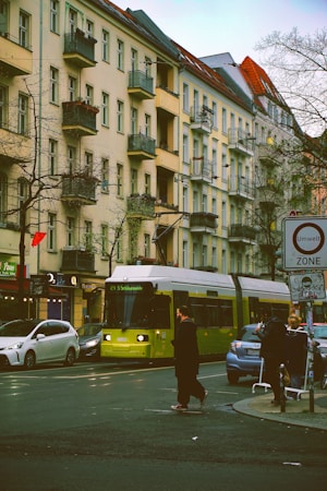 A city street with a tram in the foreground moving along the tracks. The surrounding area features tall apartment buildings with decorative balconies. Several pedestrians can be seen crossing the street, while cars wait at the intersection. The environment is urban with trees lining the sides of the road, and a street sign indicating an environmental zone.