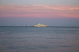 A large yacht is sailing on calm waters under a pastel-colored sky with shades of pink and blue. The horizon is dotted with a couple of small sailboats in the distance, adding depth to the serene seascape.